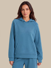 A woman modeling a matching slate blue loungewear set, featuring a pullover hoodie with a kangaroo pocket and matching sweatpants, set against a solid beige background.