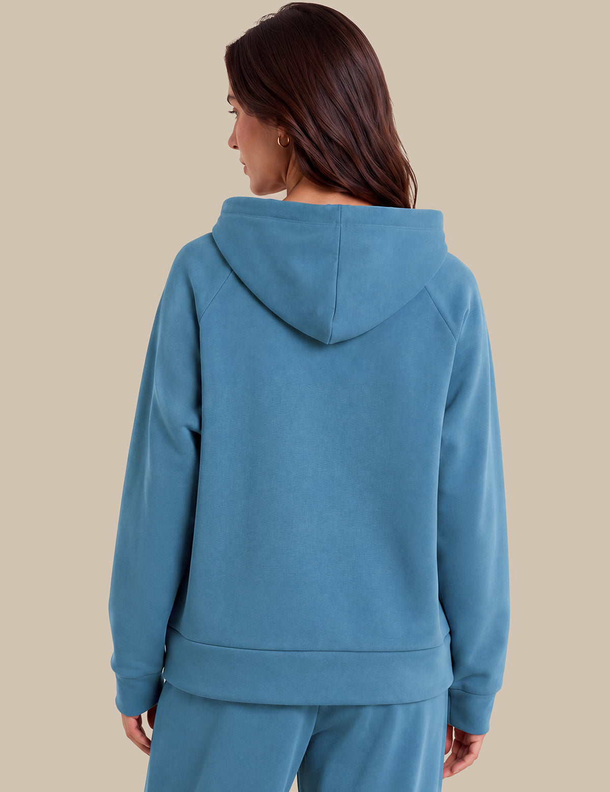A woman shown from the back wearing a dusty blue hooded sweatshirt and matching sweatpants against a solid beige background.