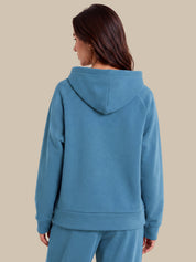 A woman shown from the back wearing a dusty blue hooded sweatshirt and matching sweatpants against a solid beige background.