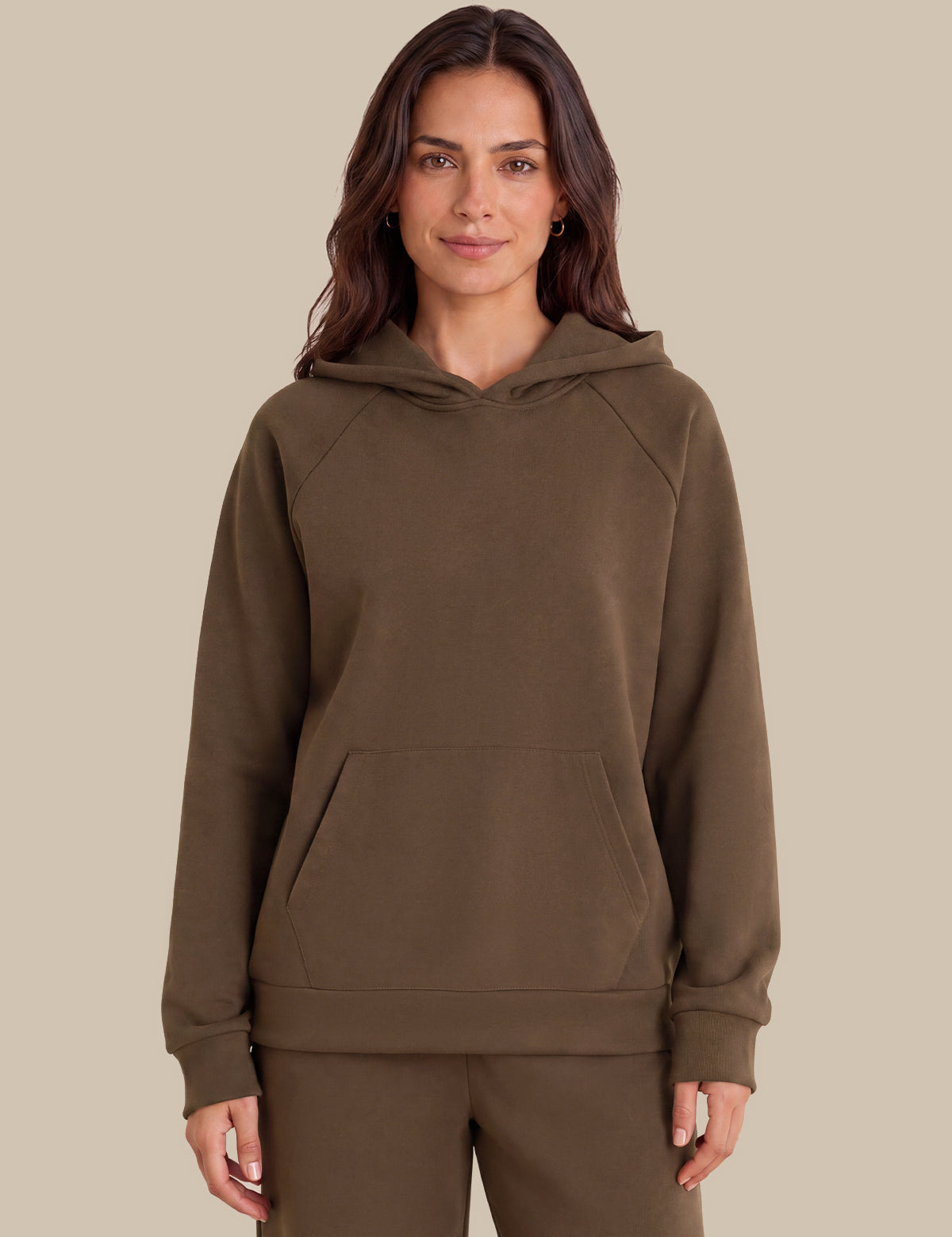A woman wearing a solid brown pullover hoodie with a kangaroo pocket and matching sweatpants, standing against a solid beige background.