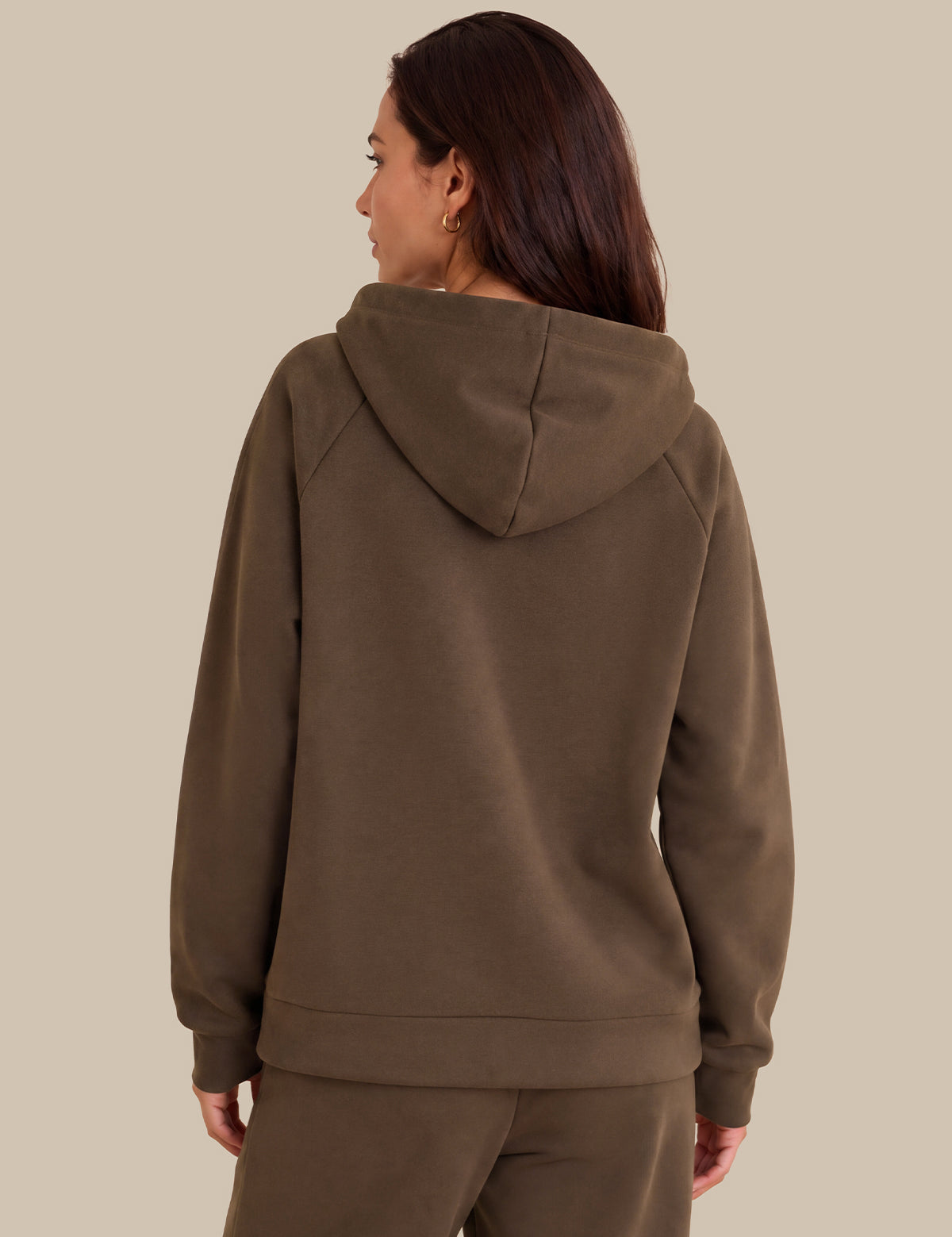 A woman shown from the back wearing a brown hooded sweatshirt and matching sweatpants against a solid beige background.