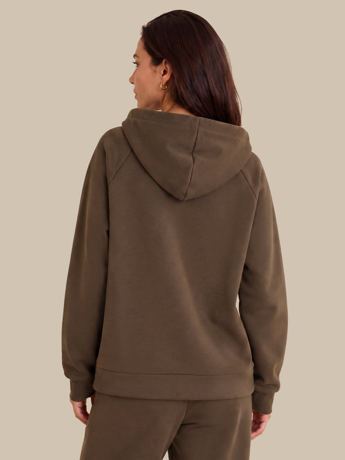 A woman shown from the back wearing a brown hooded sweatshirt and matching sweatpants against a solid beige background.