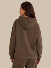 A woman shown from the back wearing a brown hooded sweatshirt and matching sweatpants against a solid beige background.