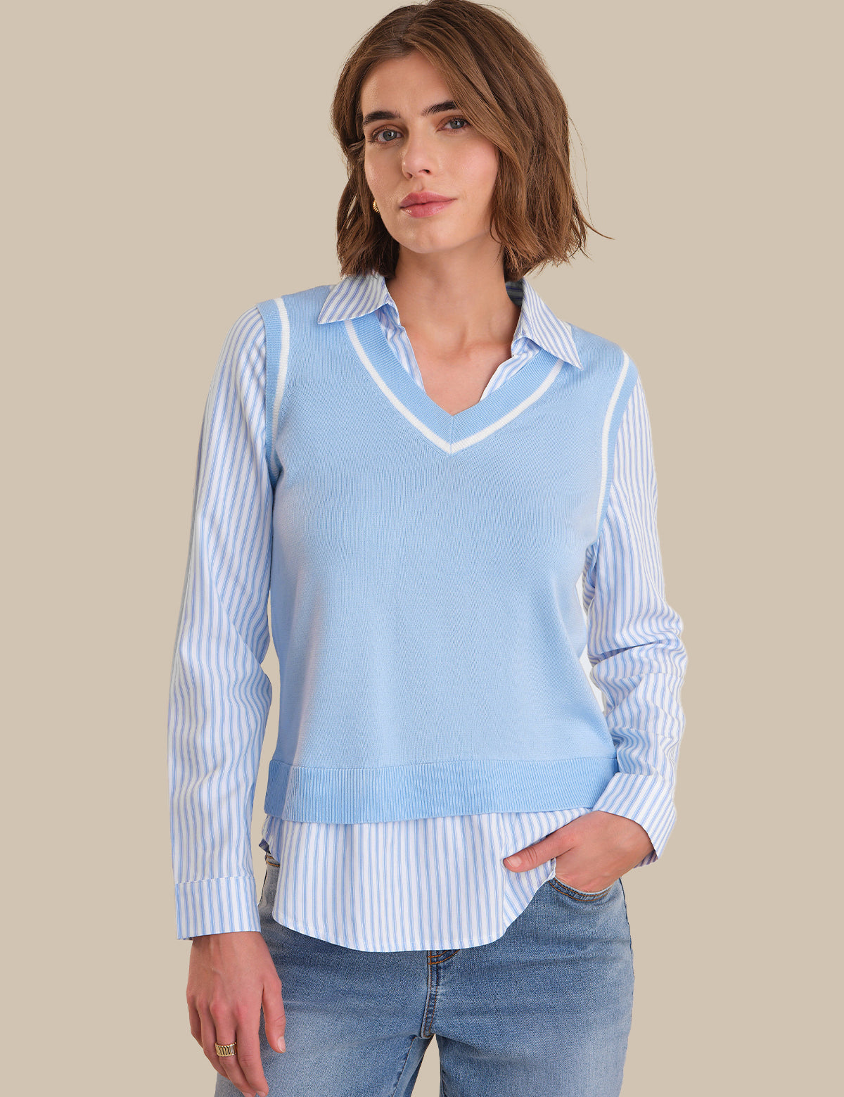 Woman wearing a light blue sweater vest over a striped shirt with jeans on a beige background