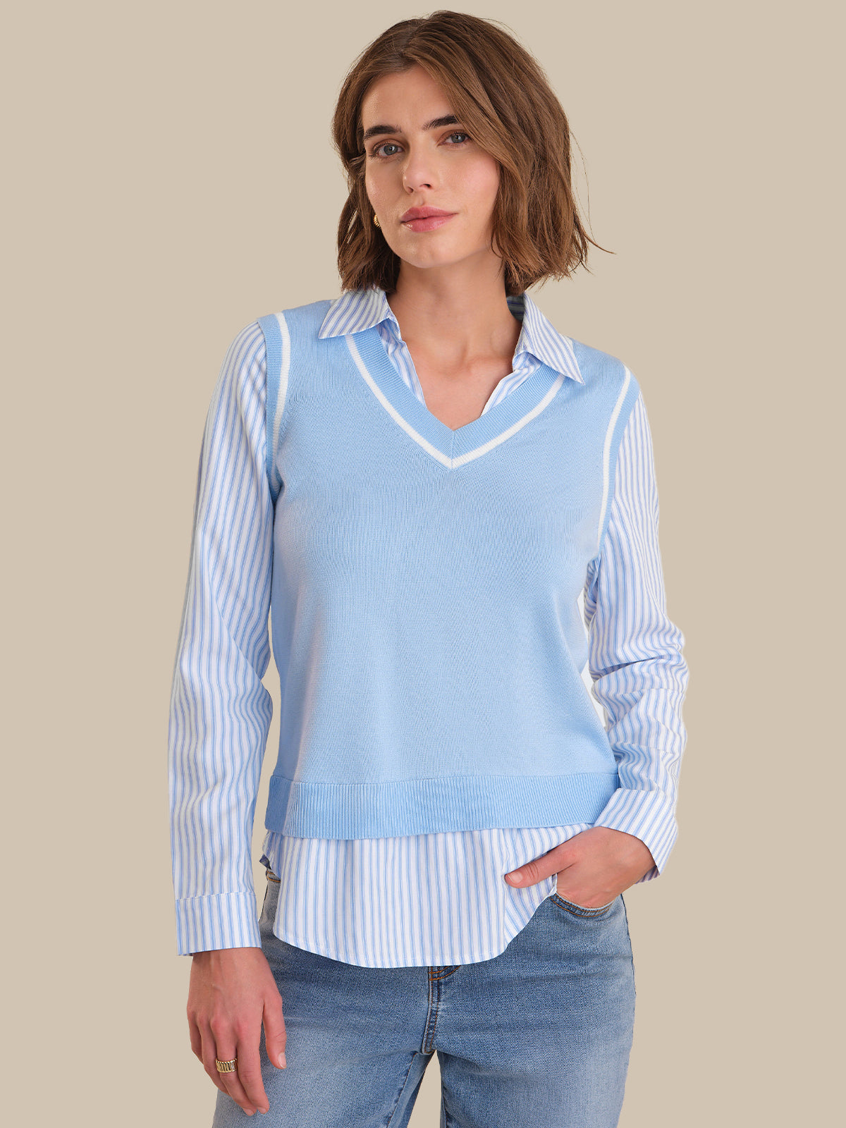 Woman wearing a light blue sweater vest over a striped shirt with jeans on a beige background