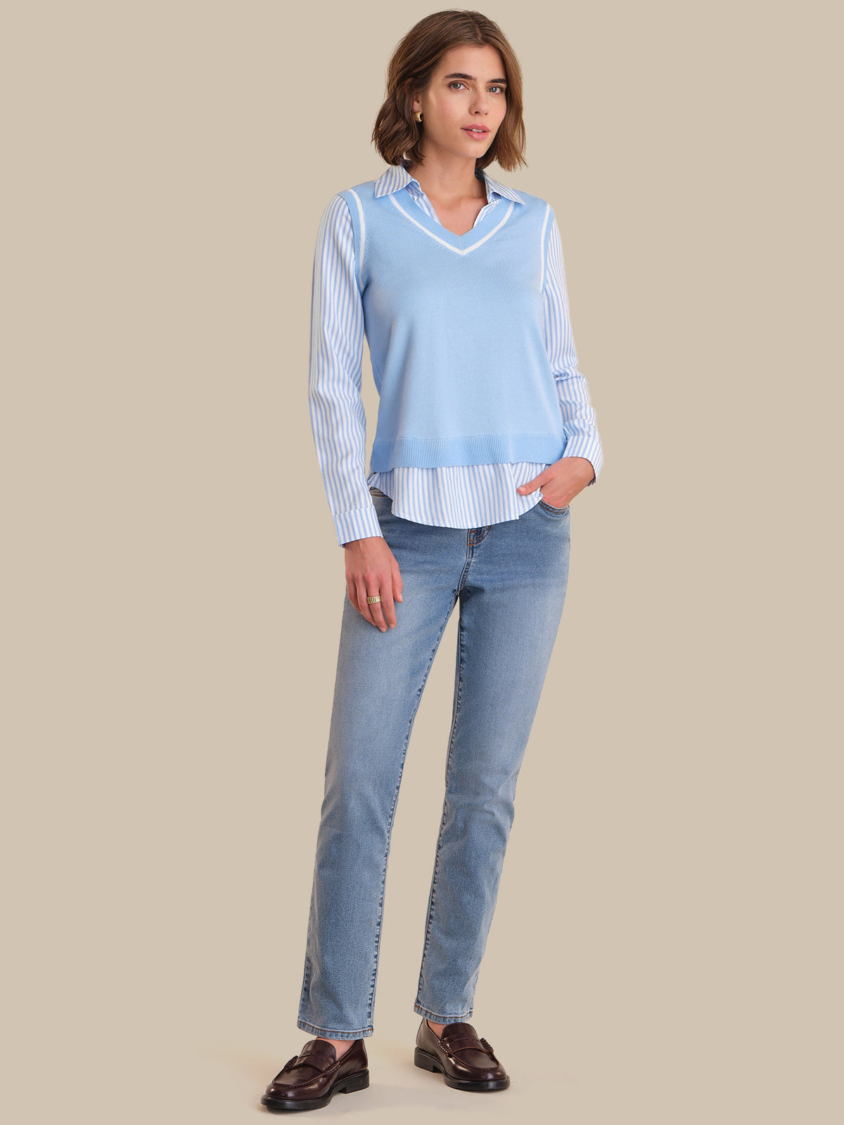 Woman wearing a light blue sweater over a striped shirt with jeans on a beige background