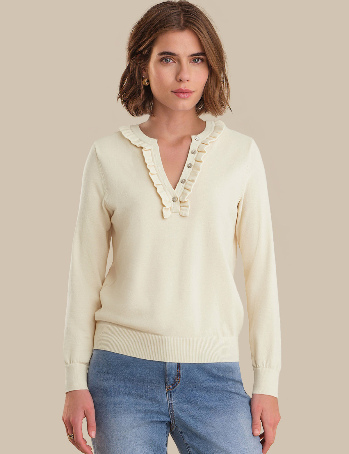 Woman wearing a cream sweater with ruffled neckline and blue jeans on a beige background