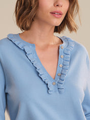 Light blue sweater with ruffled neckline and button details on a neutral background