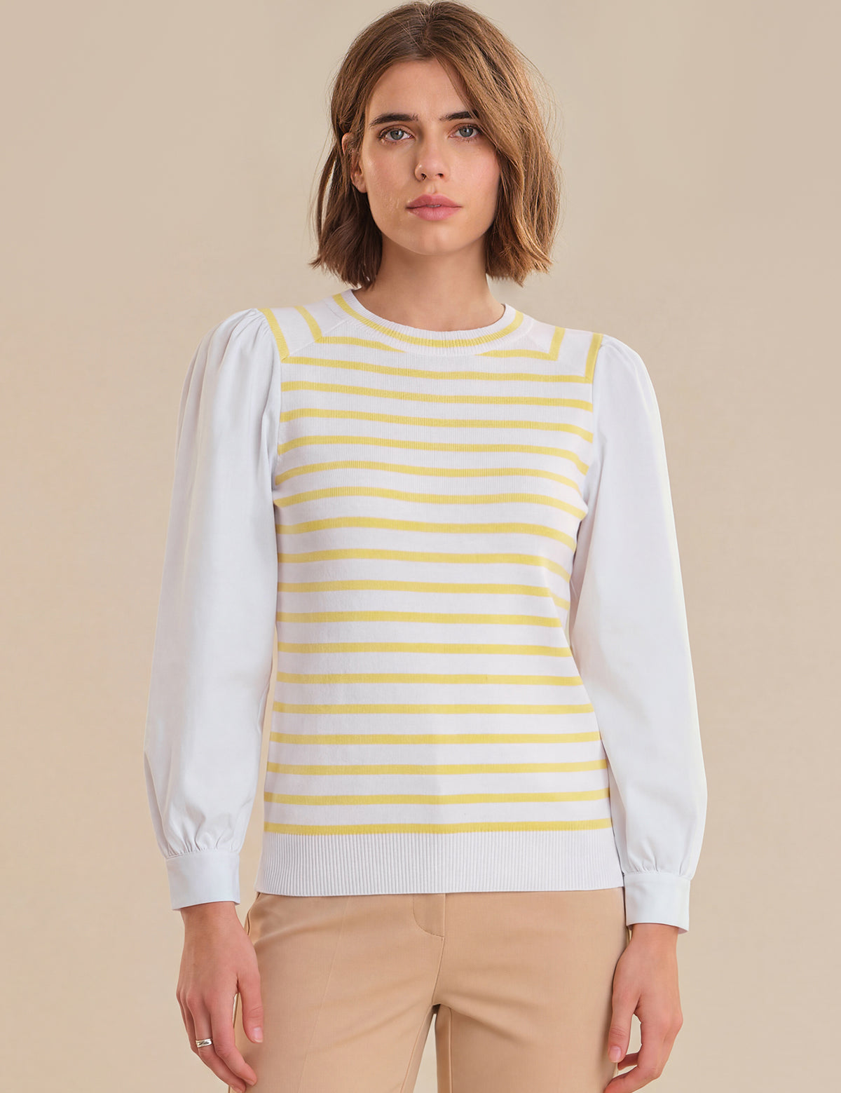 Woman wearing a white long-sleeve shirt under a yellow and white striped sweater on a beige background