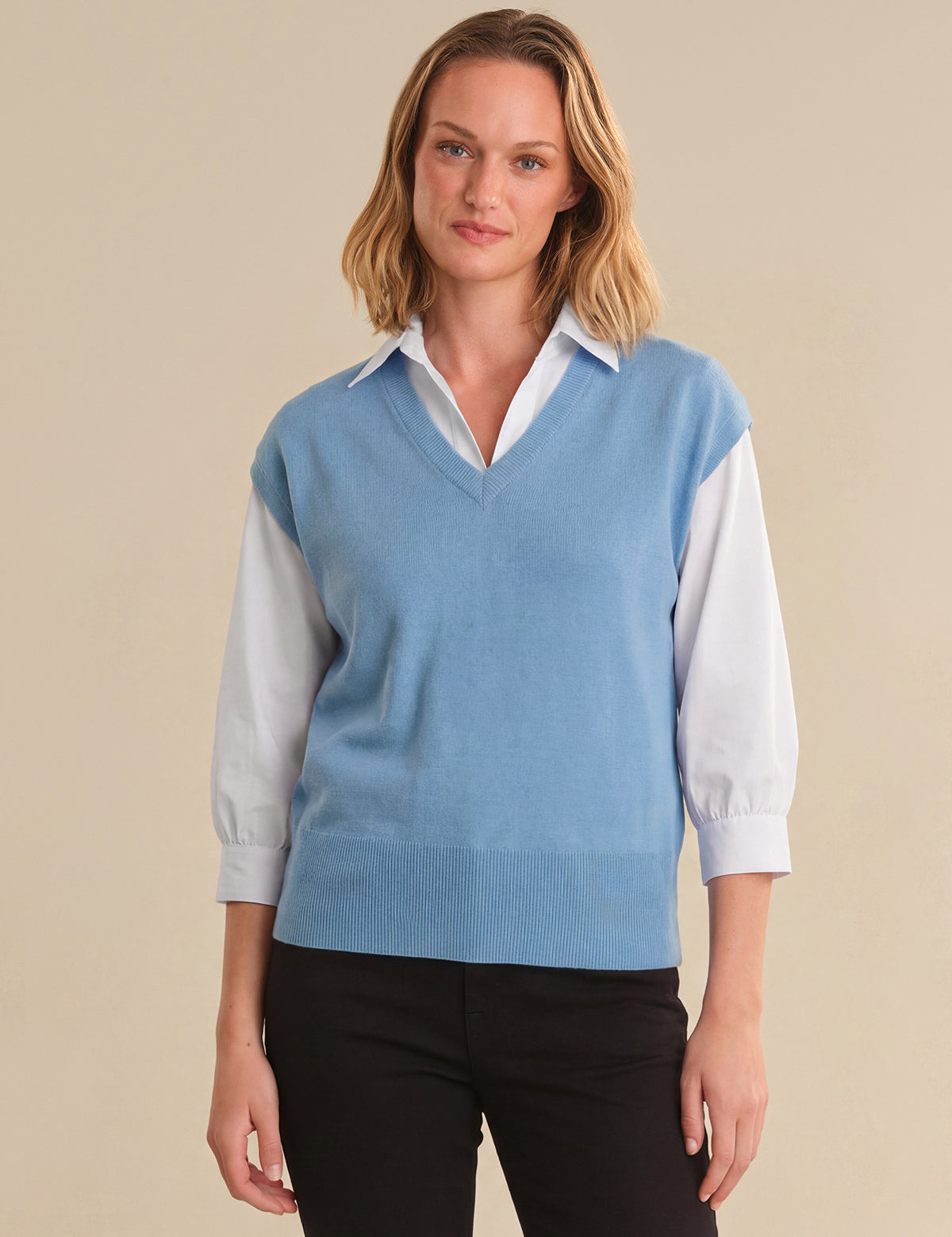 Woman wearing a light blue sweater vest over a white shirt on a beige background