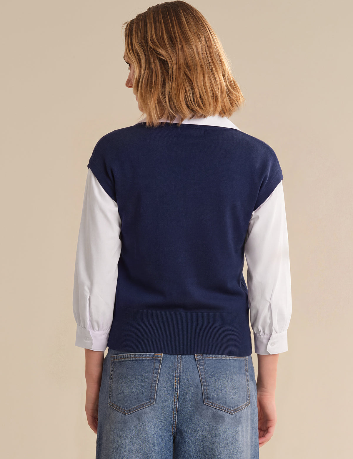 Person wearing a navy sweater over a white shirt with blue jeans on a beige background