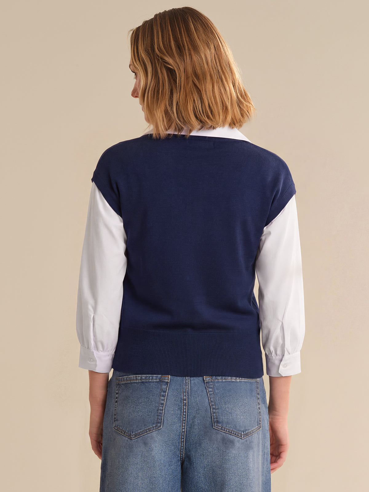 Person wearing a navy sweater over a white shirt with blue jeans on a beige background