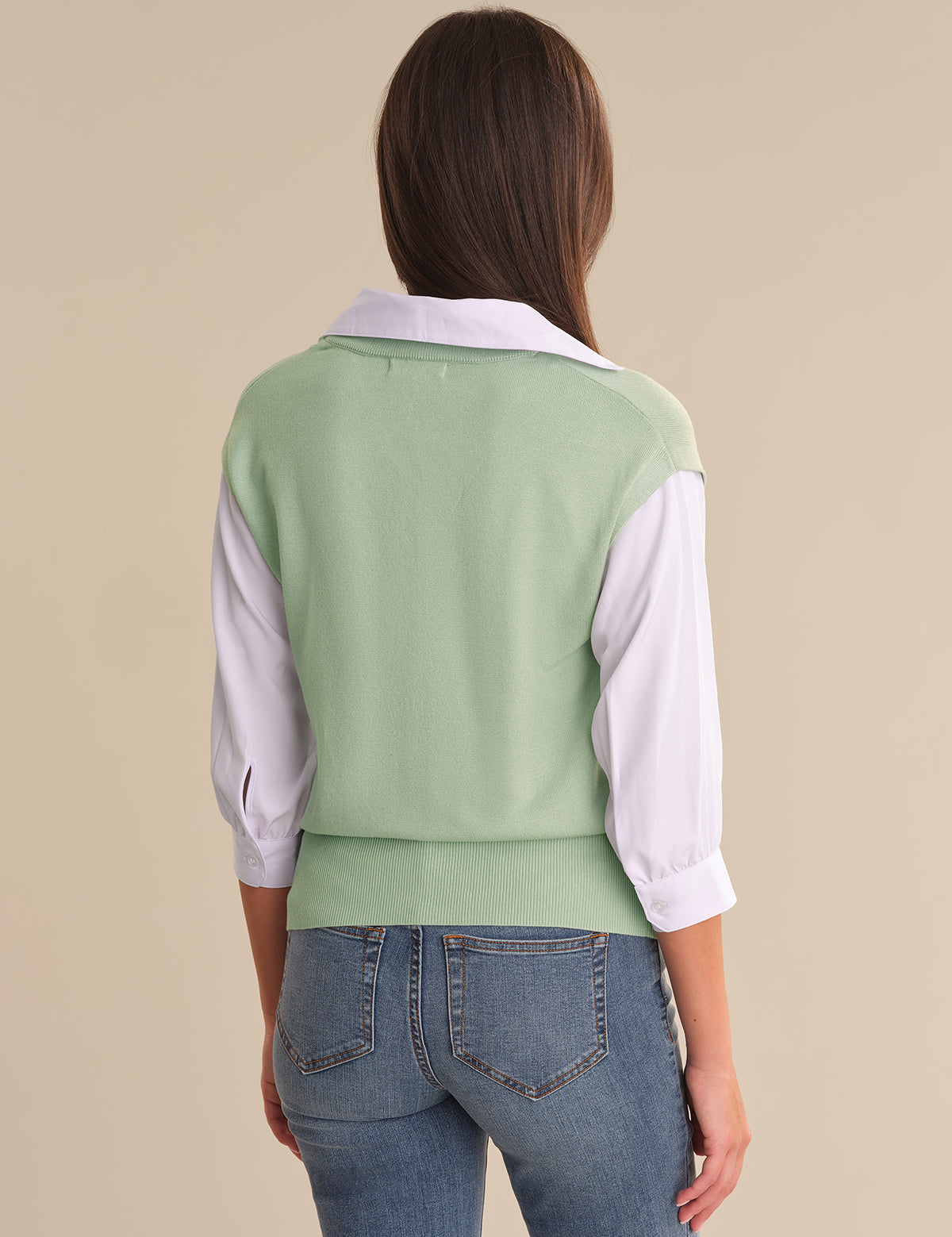 Person wearing a light green vest over a white shirt with blue jeans on a beige background