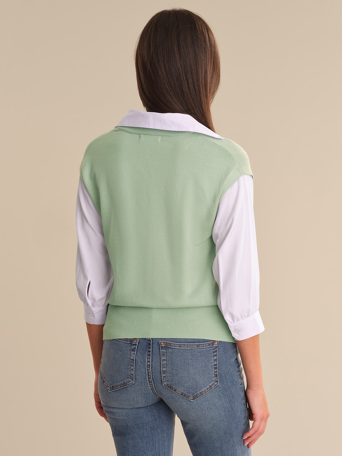 Person wearing a light green vest over a white shirt with blue jeans on a beige background