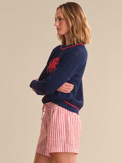 Woman wearing a navy sweater with a red design and red and white striped shorts on a beige background