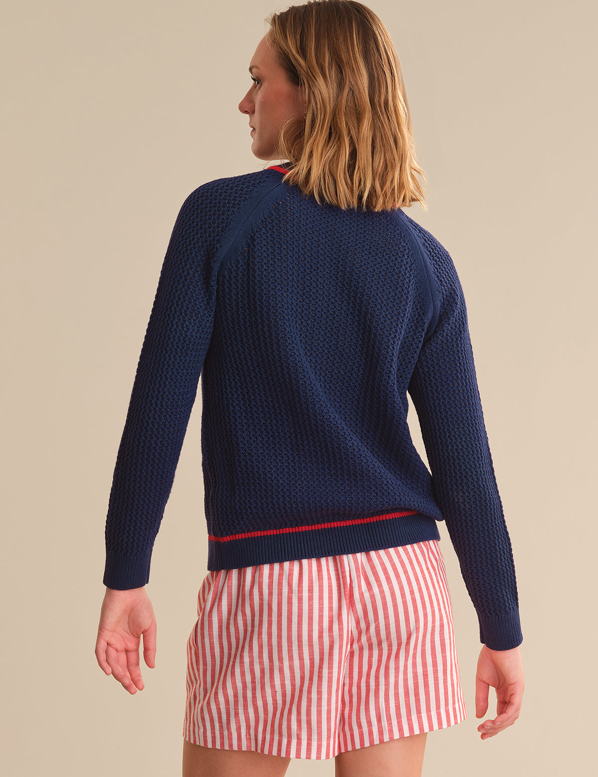 Woman wearing a navy blue knitted cardigan over a red and white striped shirt with beige shorts on a beige background.