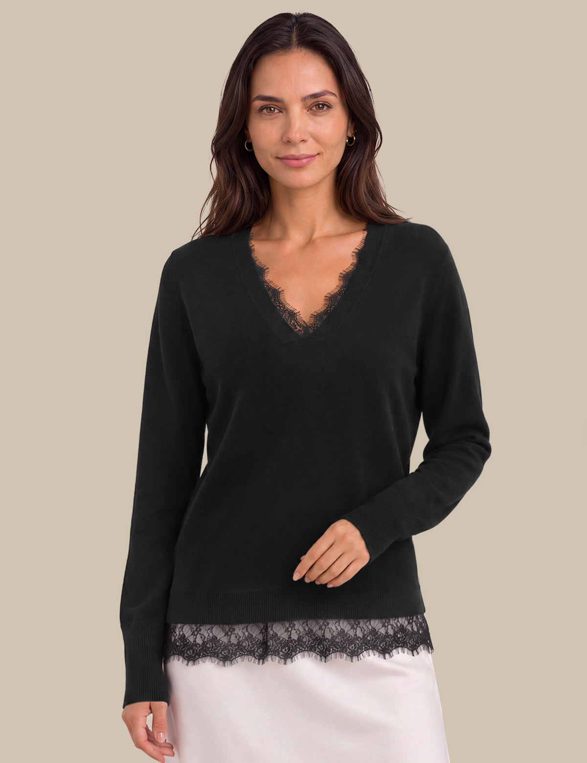 A woman modeling a black long-sleeve V-neck sweater with delicate lace trim at the neckline and hem, paired with a white skirt, against a solid beige background.