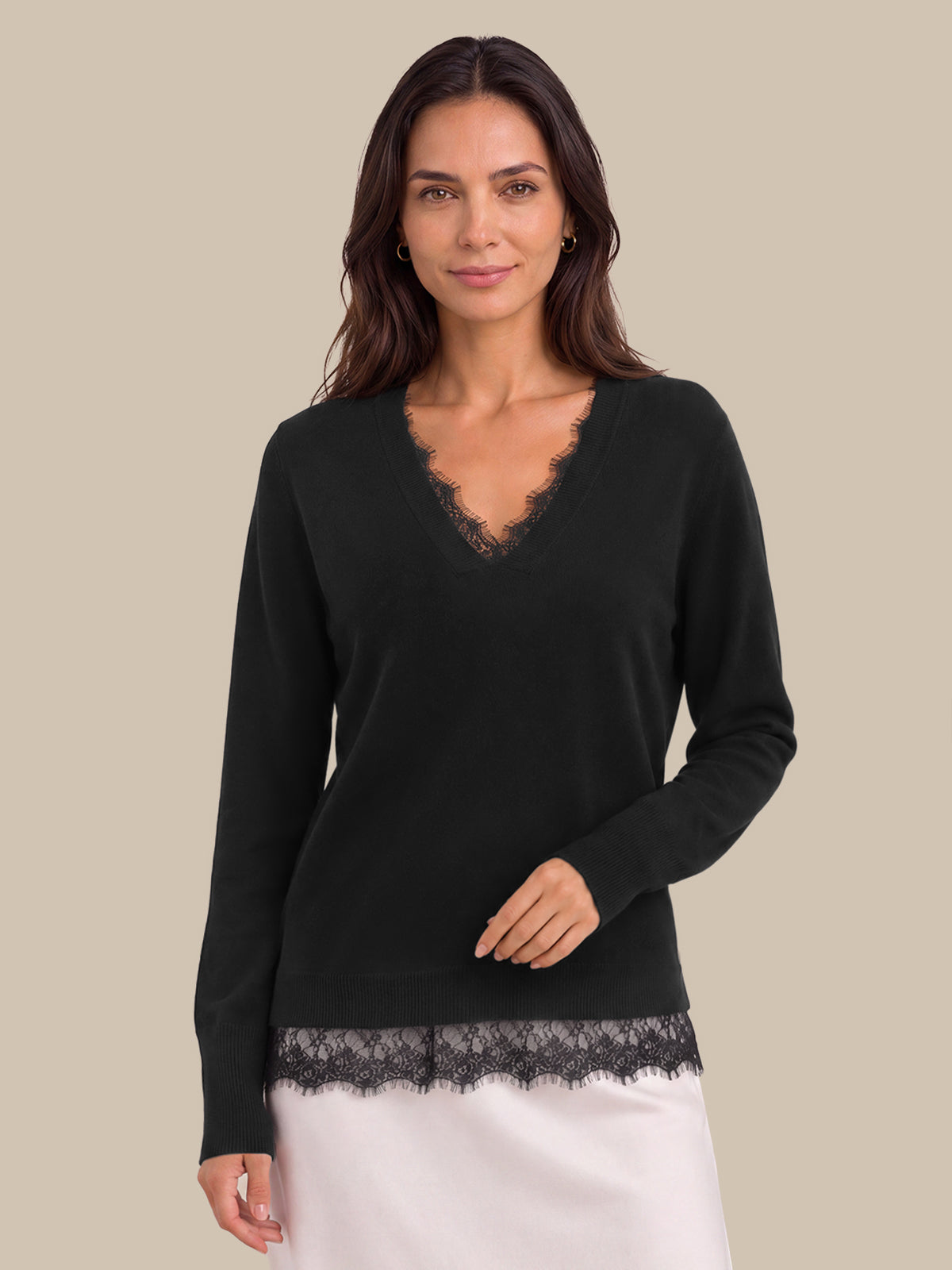 A woman modeling a black long-sleeve V-neck sweater with delicate lace trim at the neckline and hem, paired with a white skirt, against a solid beige background.