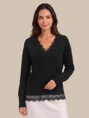 A woman modeling a black long-sleeve V-neck sweater with delicate lace trim at the neckline and hem, paired with a white skirt, against a solid beige background.