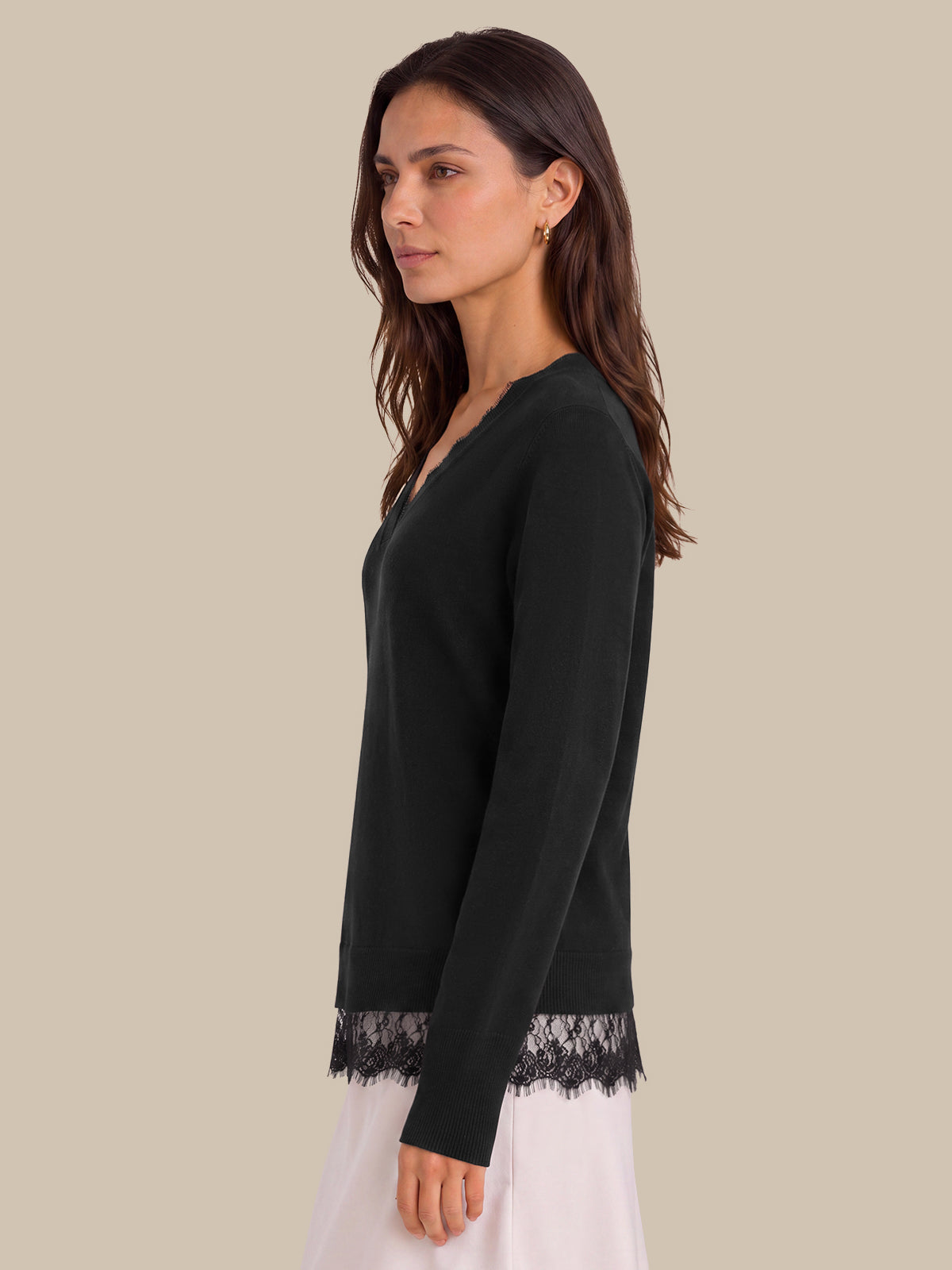 A woman shown in side profile wearing a black long-sleeve V-neck top with black lace trim at the hem and cuffs, paired with a white skirt, against a solid beige background.