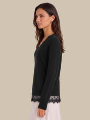 A woman shown in side profile wearing a black long-sleeve V-neck top with black lace trim at the hem and cuffs, paired with a white skirt, against a solid beige background.
