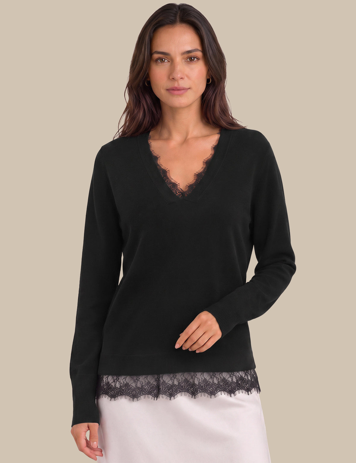 A woman modeling a black long-sleeve knit sweater with a scalloped lace V-neckline and matching lace hem, paired with a white skirt, against a solid beige background.