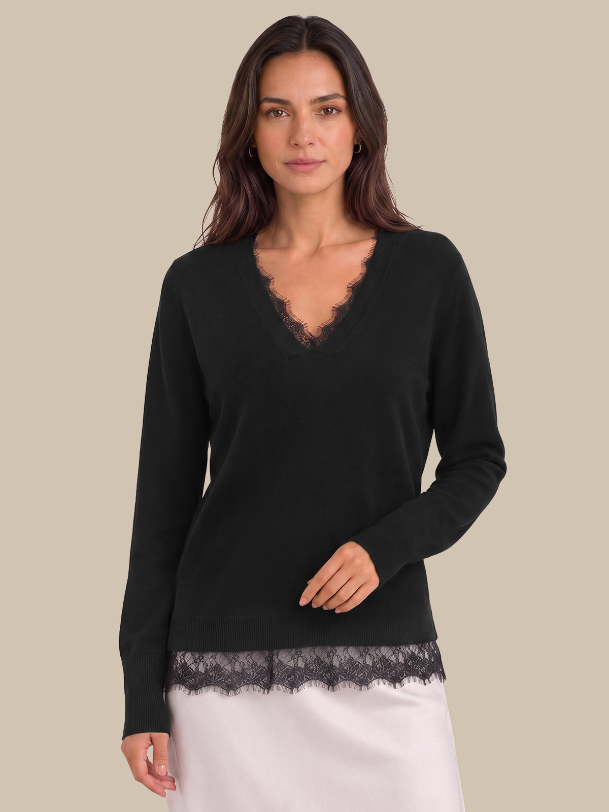 A woman modeling a black long-sleeve knit sweater with a scalloped lace V-neckline and matching lace hem, paired with a white skirt, against a solid beige background.