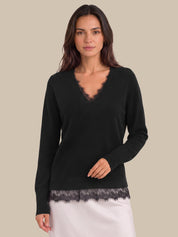 A woman modeling a black long-sleeve knit sweater with a scalloped lace V-neckline and matching lace hem, paired with a white skirt, against a solid beige background.