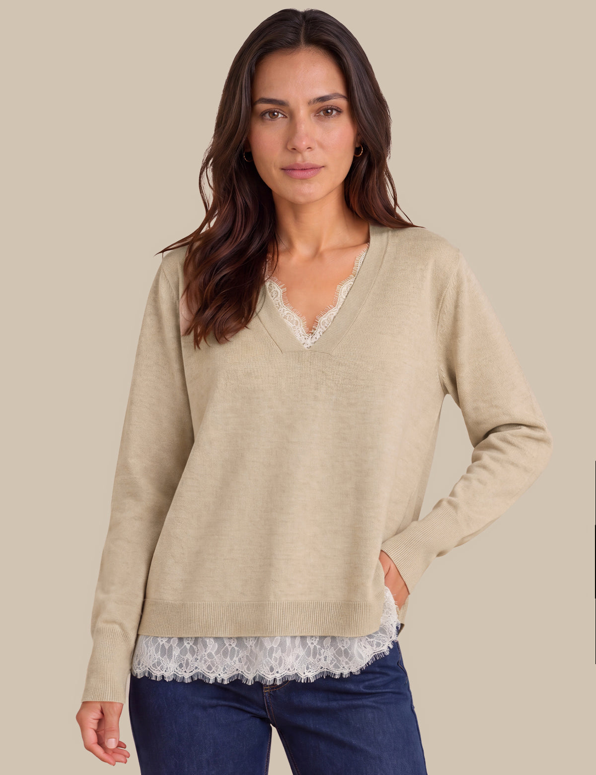 A woman wearing a beige long-sleeve V-neck sweater with a white lace trim at the neckline and hem, layered over a white lace top and paired with dark blue jeans, against a solid beige background.