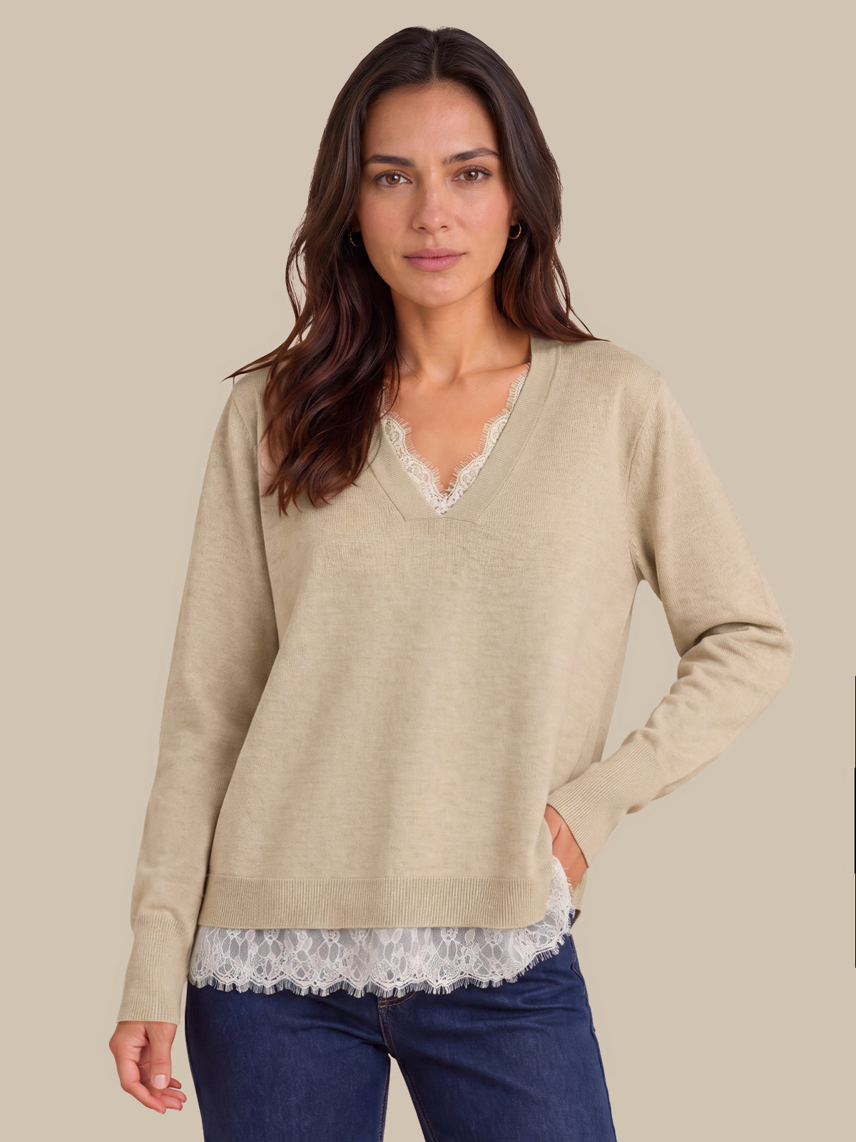 A woman wearing a beige long-sleeve V-neck sweater with a white lace trim at the neckline and hem, layered over a white lace top and paired with dark blue jeans, against a solid beige background.