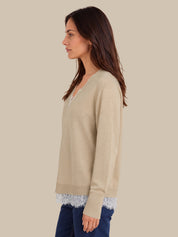 A woman shown in side profile wearing a light beige knit sweater with a V-neck and long sleeves, layered over a white lace hem, paired with dark blue jeans, against a solid beige background.