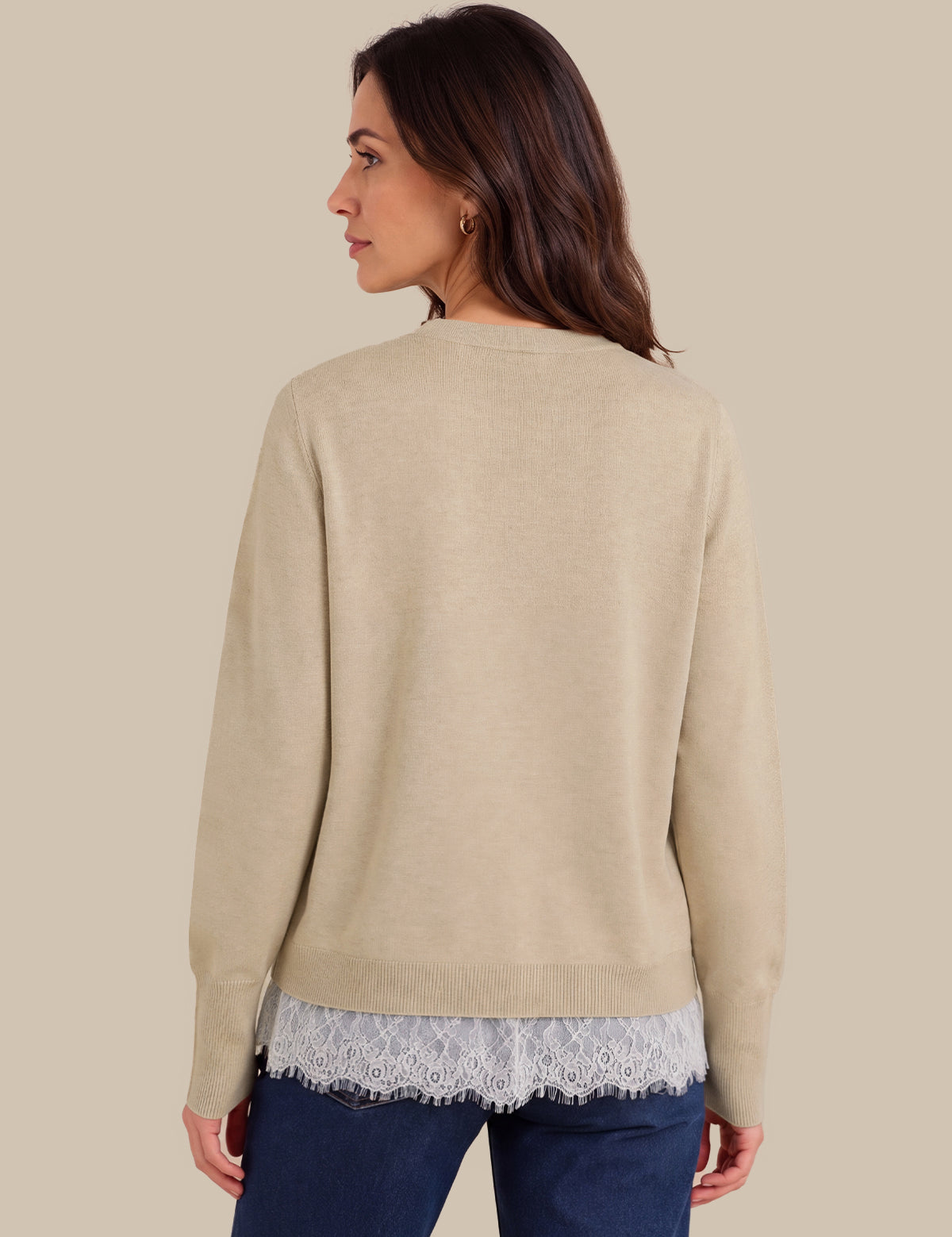 A woman shown from the back wearing a light beige knit sweater over a white lace hem top and dark blue jeans, set against a solid beige background.