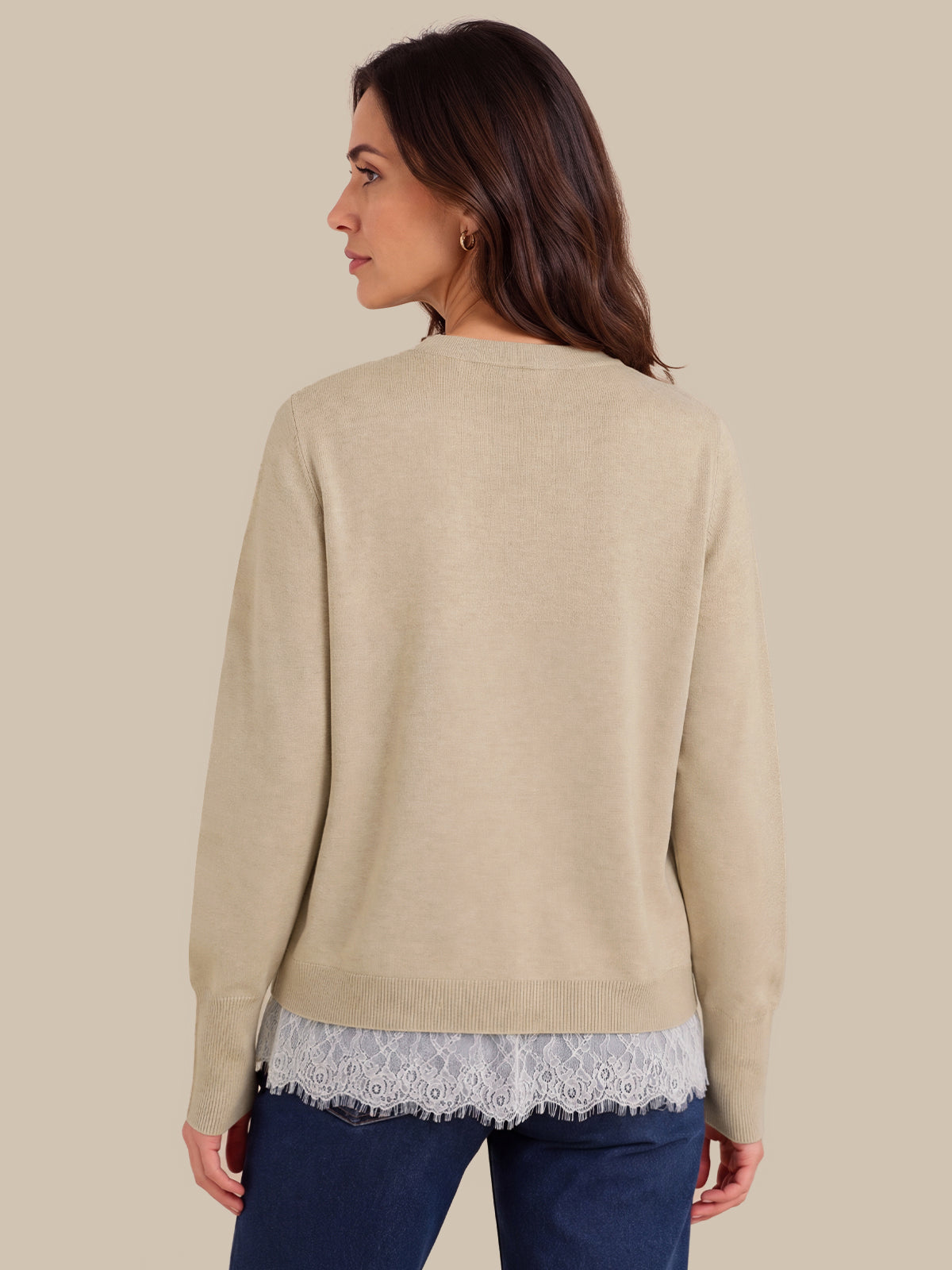 A woman shown from the back wearing a light beige knit sweater over a white lace hem top and dark blue jeans, set against a solid beige background.