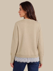 A woman shown from the back wearing a light beige knit sweater over a white lace hem top and dark blue jeans, set against a solid beige background.