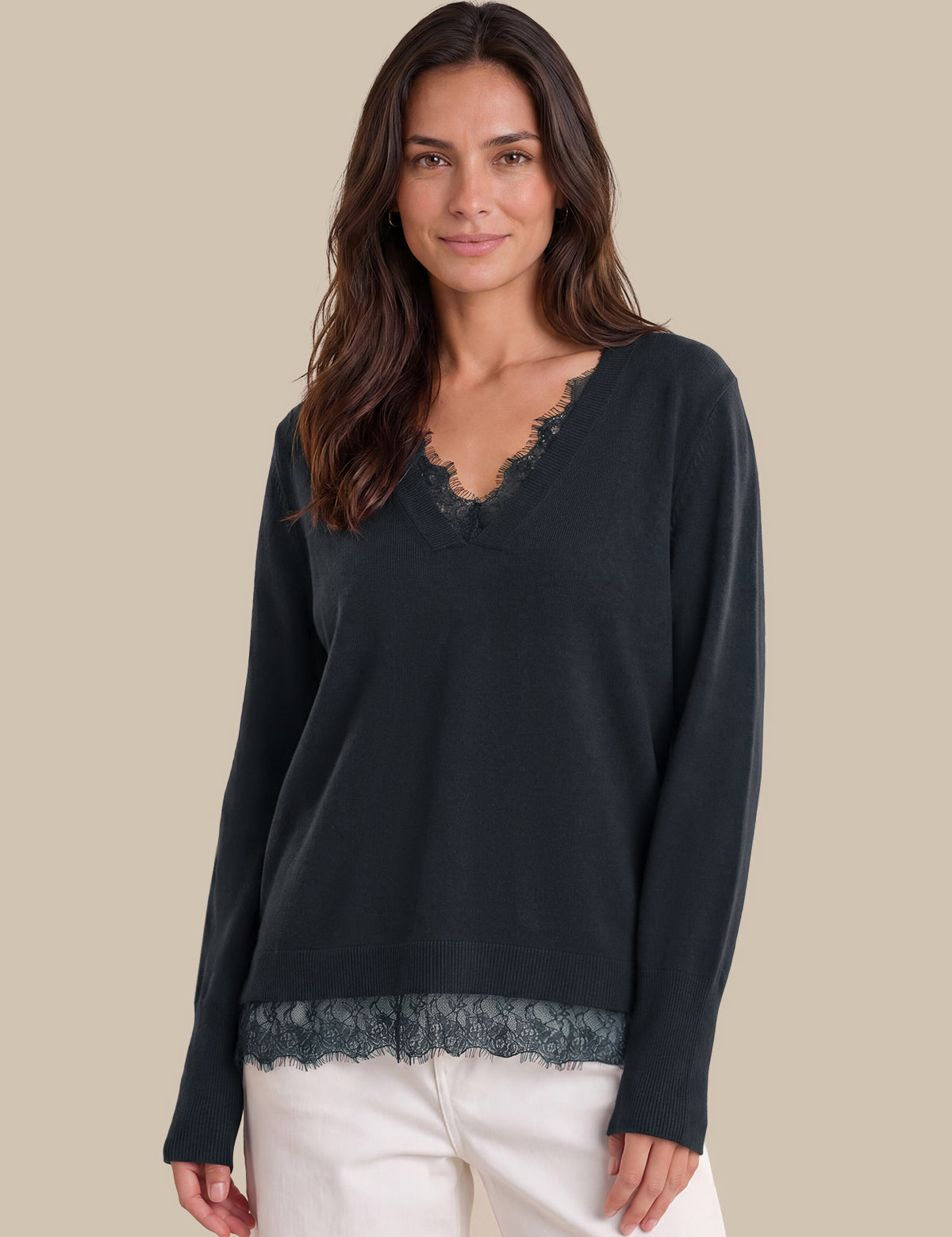A woman modeling a dark blue long-sleeve knit top with a V-neck and lace trim, paired with white pants, against a solid beige background.