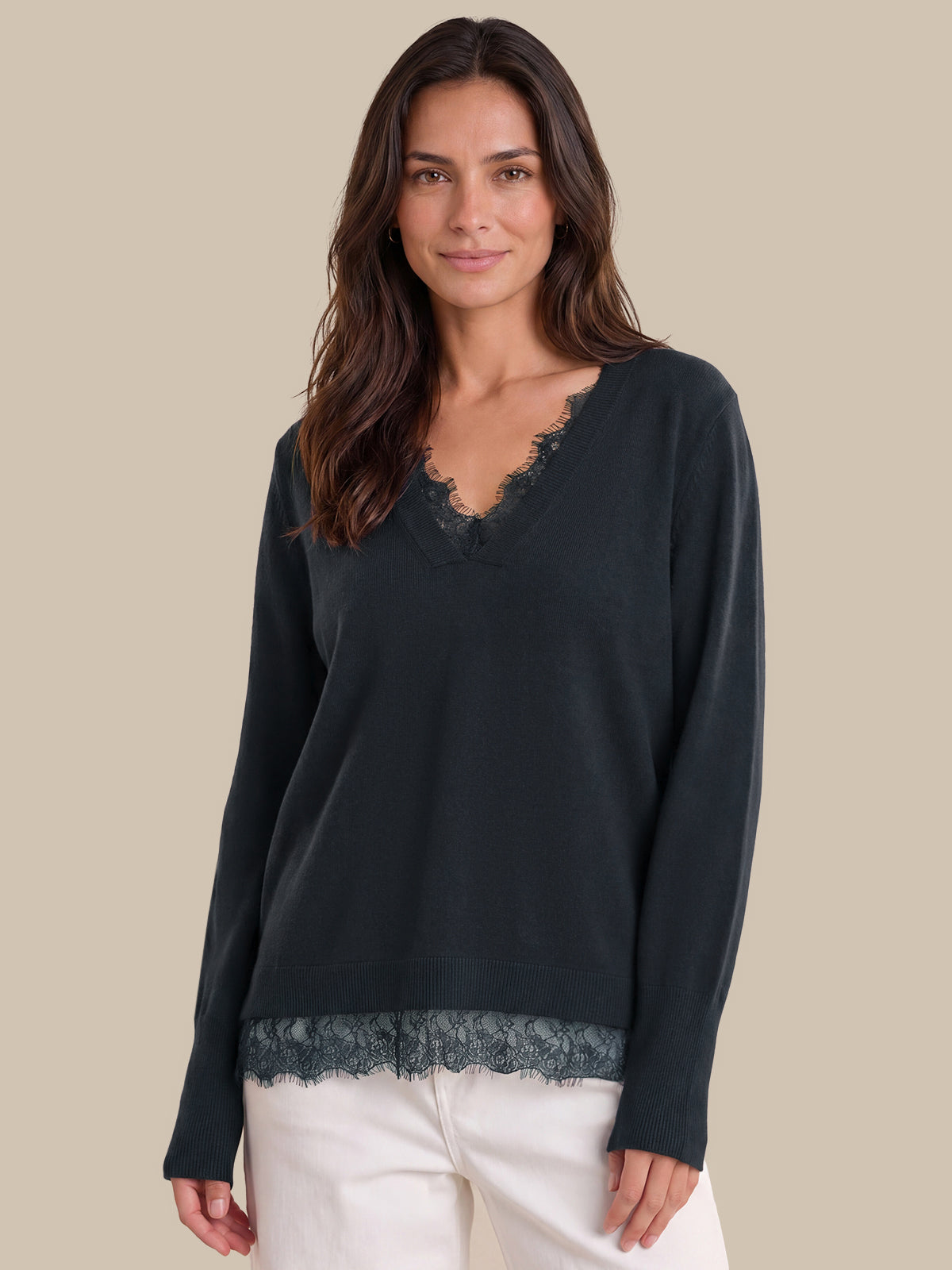 A woman modeling a dark blue long-sleeve knit top with a V-neck and lace trim, paired with white pants, against a solid beige background.