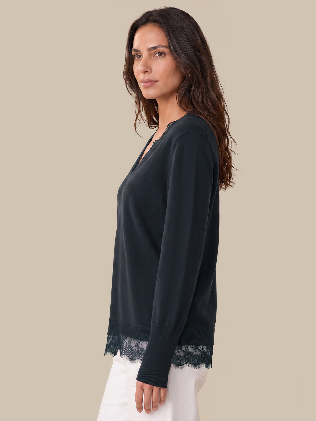 A woman shown in side profile wearing a dark blue long-sleeve top with a V-neck and lace trim at the hem, paired with white pants, against a solid beige studio background.