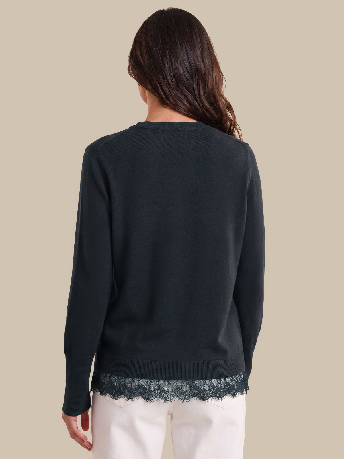 A woman shown from the back wearing a dark blue long-sleeve top with a lace hem detail, paired with white pants, against a solid beige background.