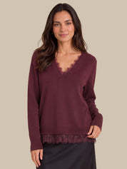 A woman wearing a long-sleeve burgundy knit sweater with a lace V-neck trim and matching lace hem, paired with a dark skirt, against a solid beige background.