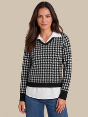 A woman modeling a black and white houndstooth knit sweater with a white collar and black trim, paired with blue jeans, against a solid beige background.