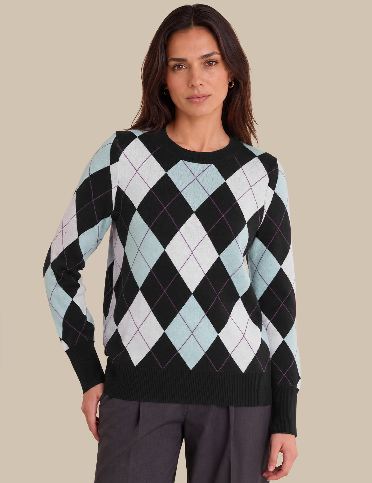 A woman modeling a long-sleeve crewneck sweater with an argyle pattern in black, white, and light blue, paired with dark gray trousers, against a solid beige background.