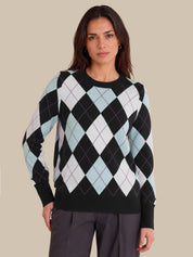 A woman modeling a long-sleeve crewneck sweater with an argyle pattern in black, white, and light blue, paired with dark gray trousers, against a solid beige background.