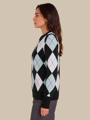 A woman shown in side profile wearing a black knit sweater with a light blue, white, and purple argyle pattern, paired with dark gray trousers, against a solid beige background.