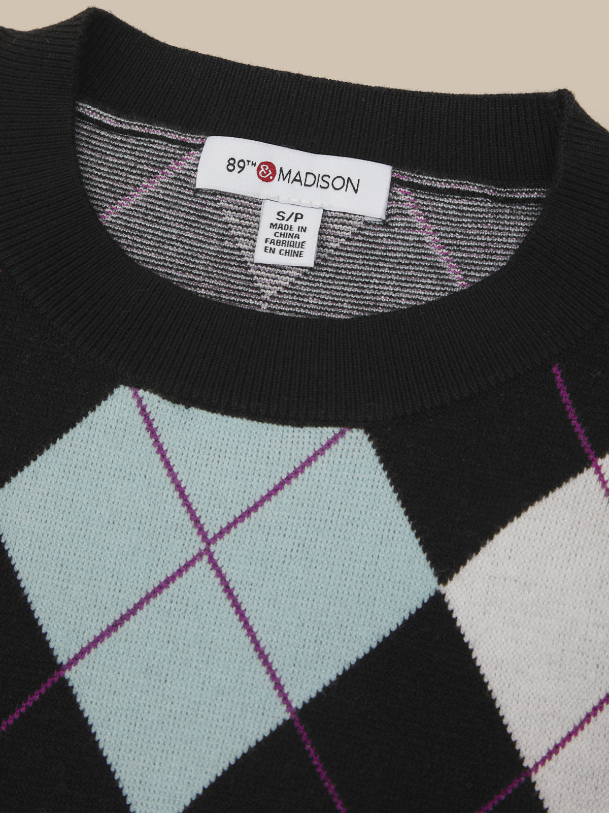 Close-up of a knit sweater with a black ribbed crew neck and an argyle pattern featuring light blue, white, and purple diamonds. The brand label '89th & MADISON' is visible inside the collar.
