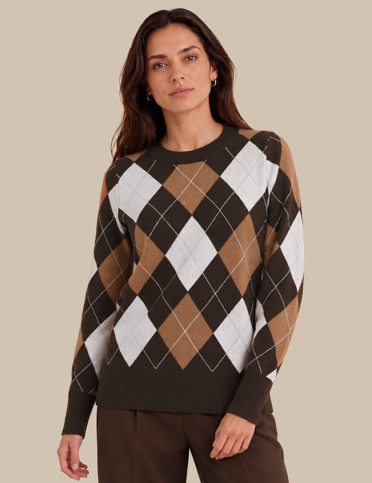 A woman wearing a long-sleeve crewneck sweater with an argyle pattern in dark brown, tan, and white, paired with dark brown trousers, set against a solid beige background.
