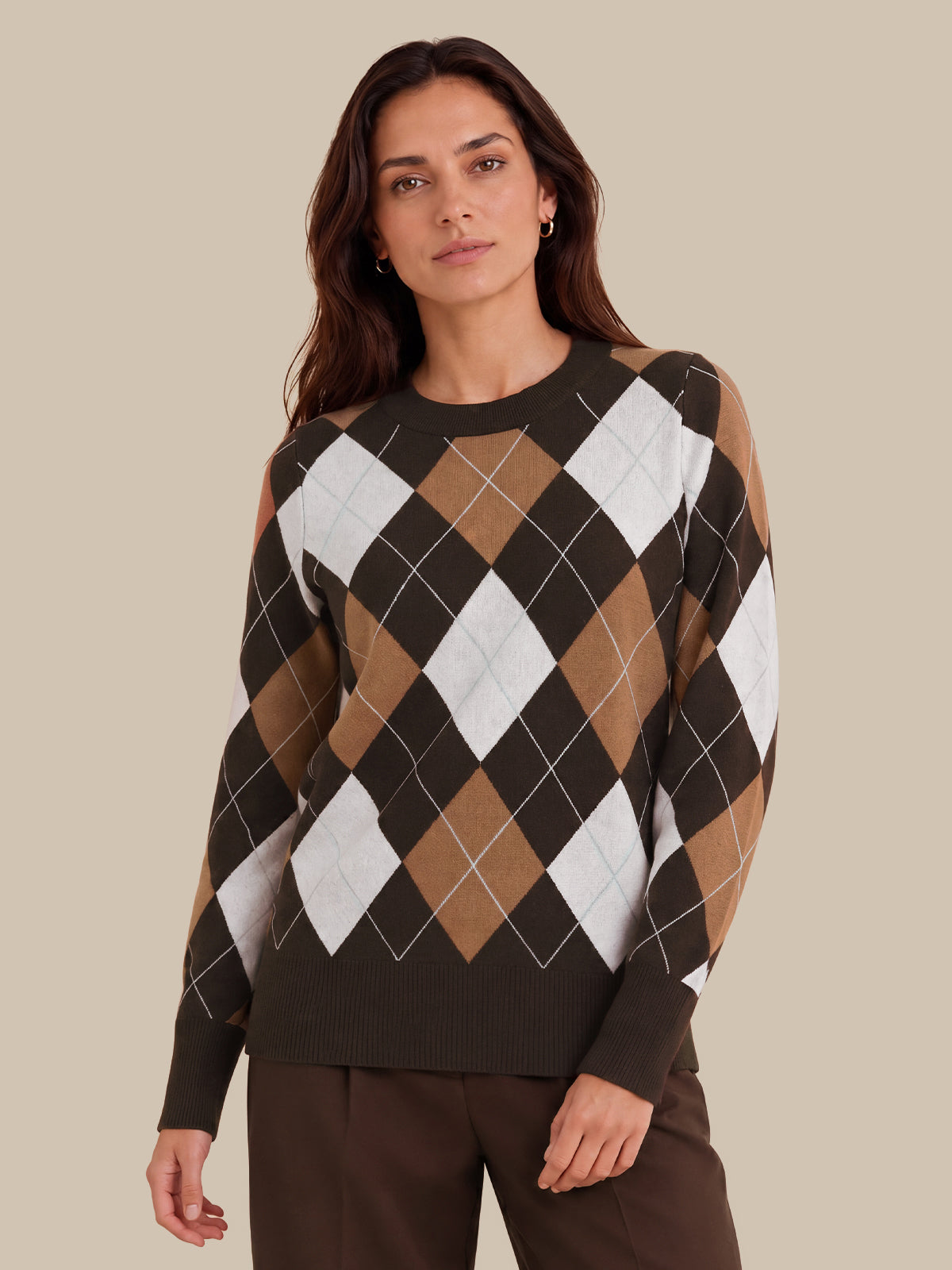 A woman wearing a long-sleeve crewneck sweater with an argyle pattern in dark brown, tan, and white, paired with dark brown trousers, set against a solid beige background.