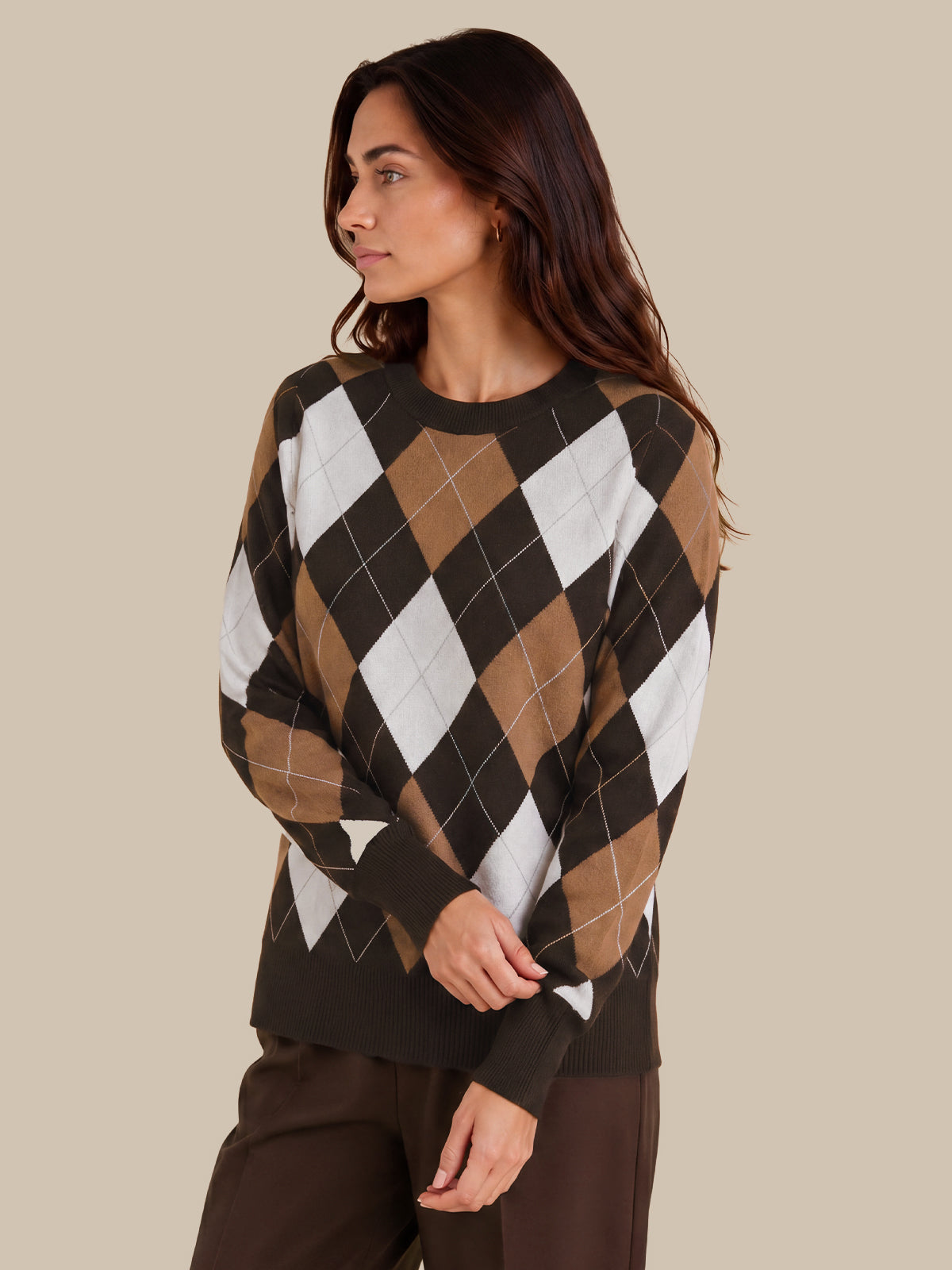 A woman wearing a long-sleeve crewneck sweater with an argyle pattern in brown, tan, white, and black, paired with dark brown pants, set against a solid beige background.