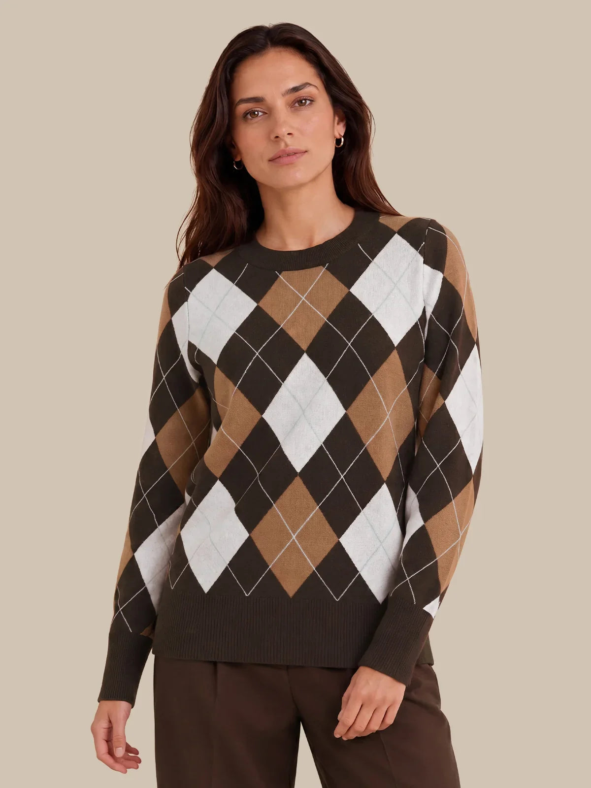 A woman wearing a long-sleeve crewneck sweater with an argyle pattern in dark brown, tan, and white, paired with dark brown trousers, set against a solid beige background.
