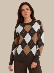 A woman wearing a long-sleeve crewneck sweater with an argyle pattern in dark brown, tan, and white, paired with dark brown trousers, set against a solid beige background.