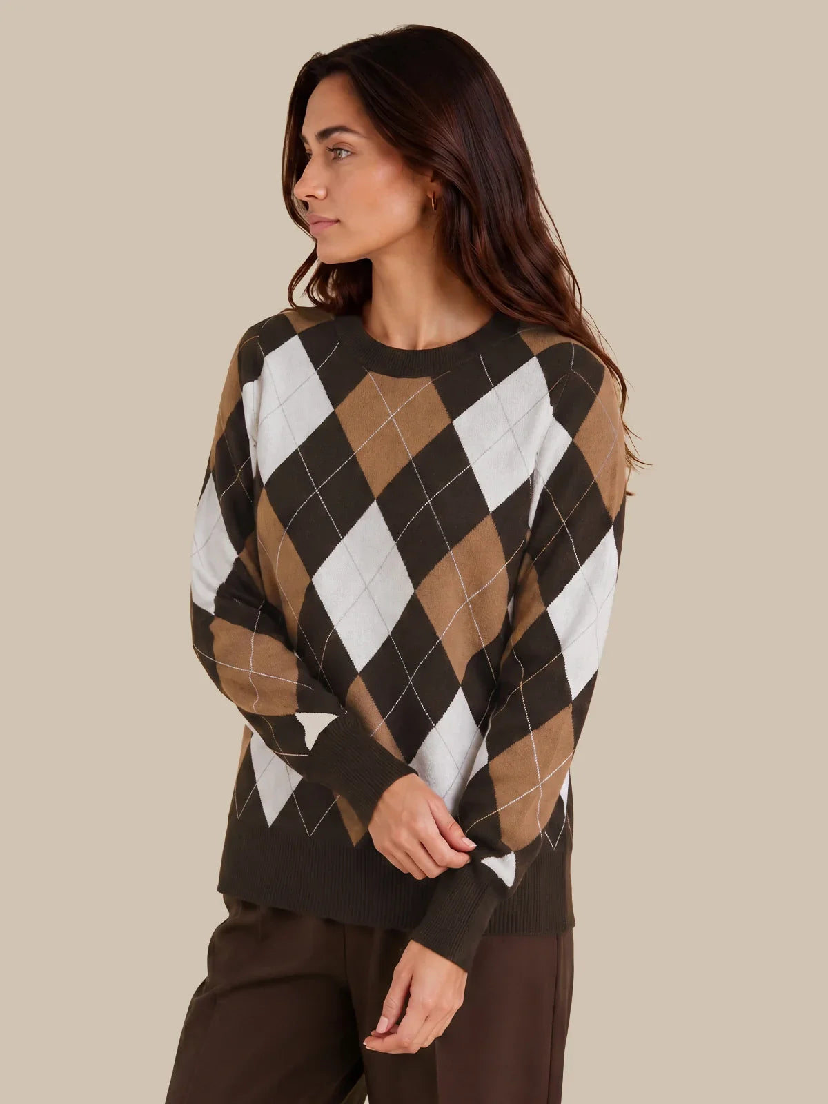 A woman wearing a long-sleeve argyle sweater in brown, tan, white, and dark brown, paired with dark brown pants, set against a solid beige background.